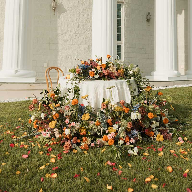 image of an abundance of flowers draped over a white tableclothed table outside in orange, red, yellow, and peach color with greenery