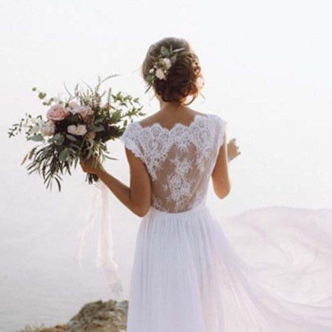 a bride facing the water wearing a white lace wedding dress with the wind blowing her train and holding a large bouquet of pink and white flowers with greenery