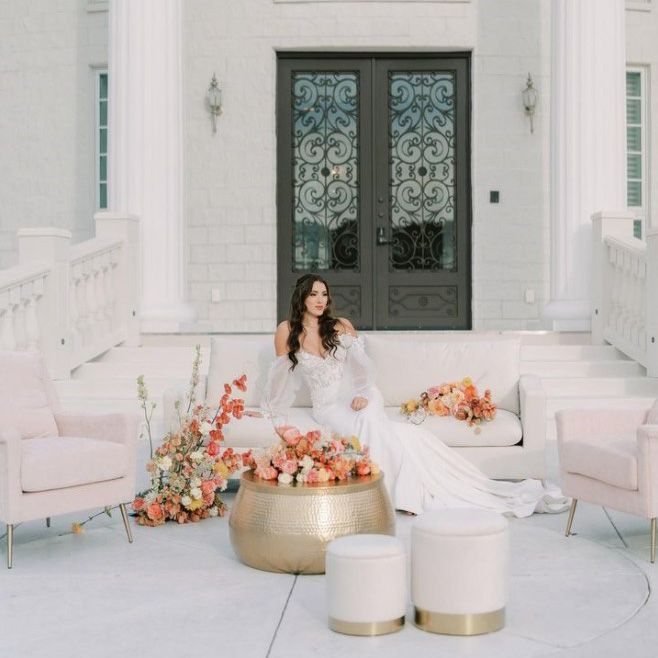 image of a bride leaning against a white ottoman with two light pink side chairs and a hammered gold coffee table strewn with light orange, pink, and white flowers with an ornate iron doors in the background