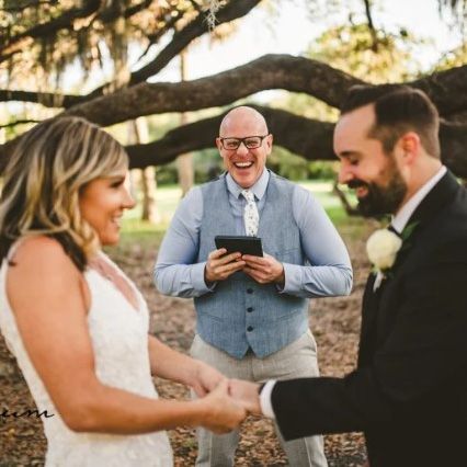 Image of a bride in a white dress and groom in a tuxedo getting married under a large tree holding hands with a wedding officiant wearing a blue vest and they are all laughing