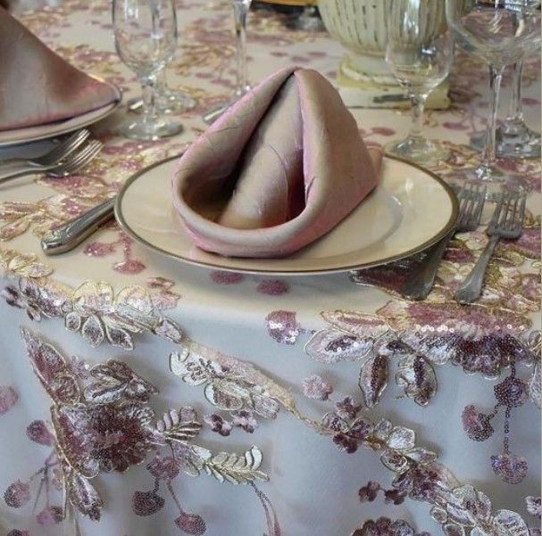 image of a table with an elaborate tablecloth with purple and gold applique flowers and formal white china, silverware and glasses with a neatly folded purple and gold tinted napkin