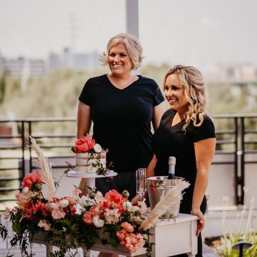 two smiling women with blonde hair both wearing all black standing in front of a table with a wedding cake and champagne in a bucket with a lot of flowers in shades of pink, white and peach with a skyline view in the background