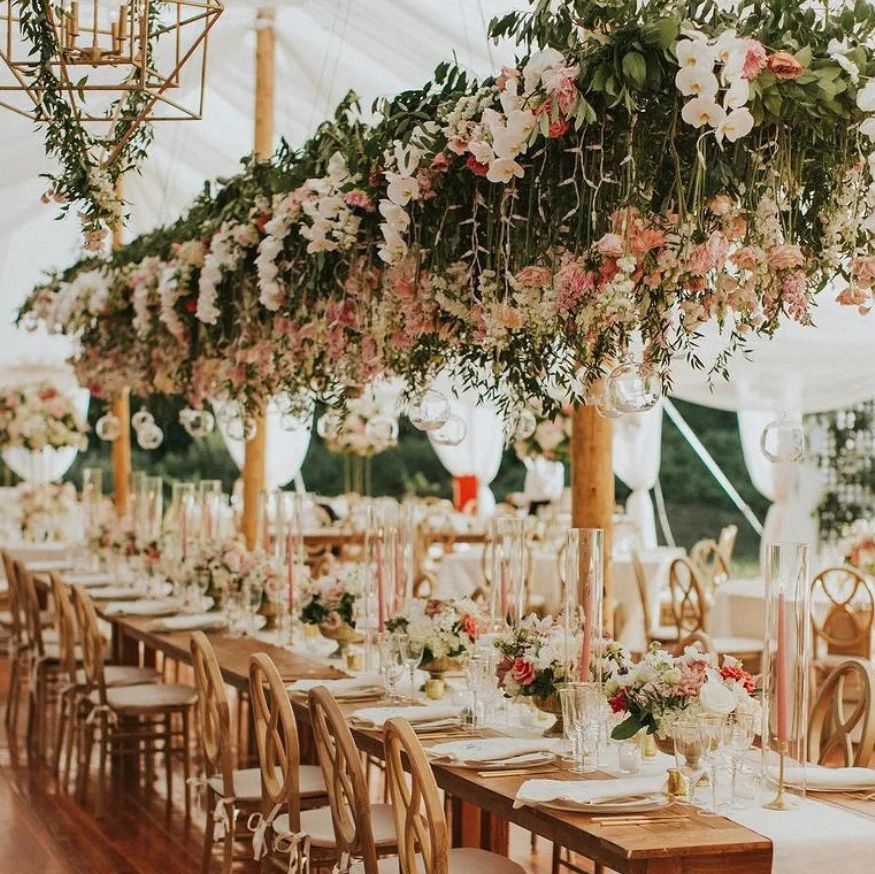 image of a long table set shabby chic place settings, gold bamboo back chairs, and a lot of flowers and greenery and chandeliers hanging from above in a white tent