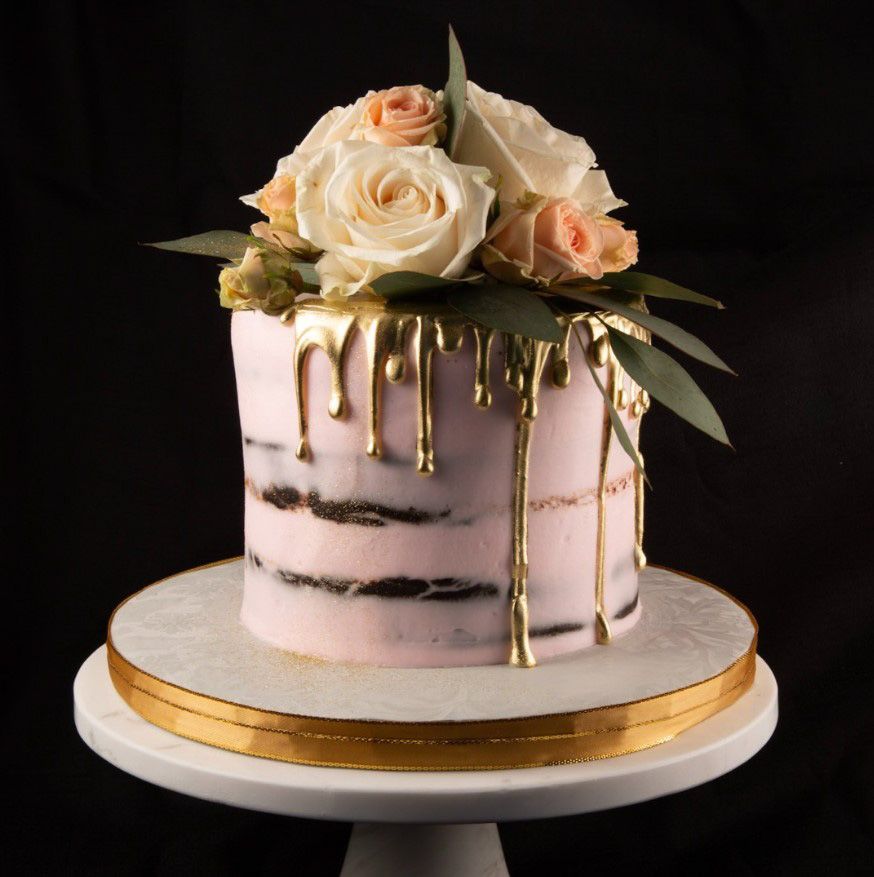 image of a cake frosted with pink icing with gold icing dripping down the sides with white and pink roses on top sitting on a cake stand with a black background
