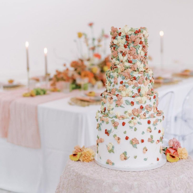 image of a 4-tiered wedding cake frosted with pink and white flowers in varying shades with light green and blue leaves set upon a small table covered in pink lace with a formal table set with candles in the background