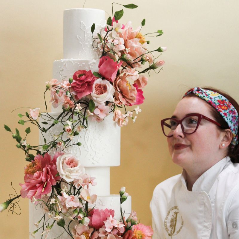 white 6-tier wedding cake with many large flowers in various colors of pink with greenery winding around the tiers with a lace applique look created in the icing with Jordan Ellen standing nearby