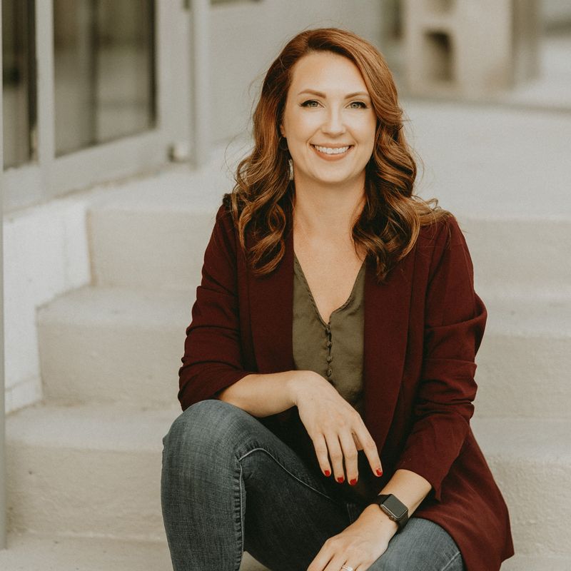 smiling woman with red hair and and dark red blazer wearing an olive green shirt and black jeans while sitting on stone steps