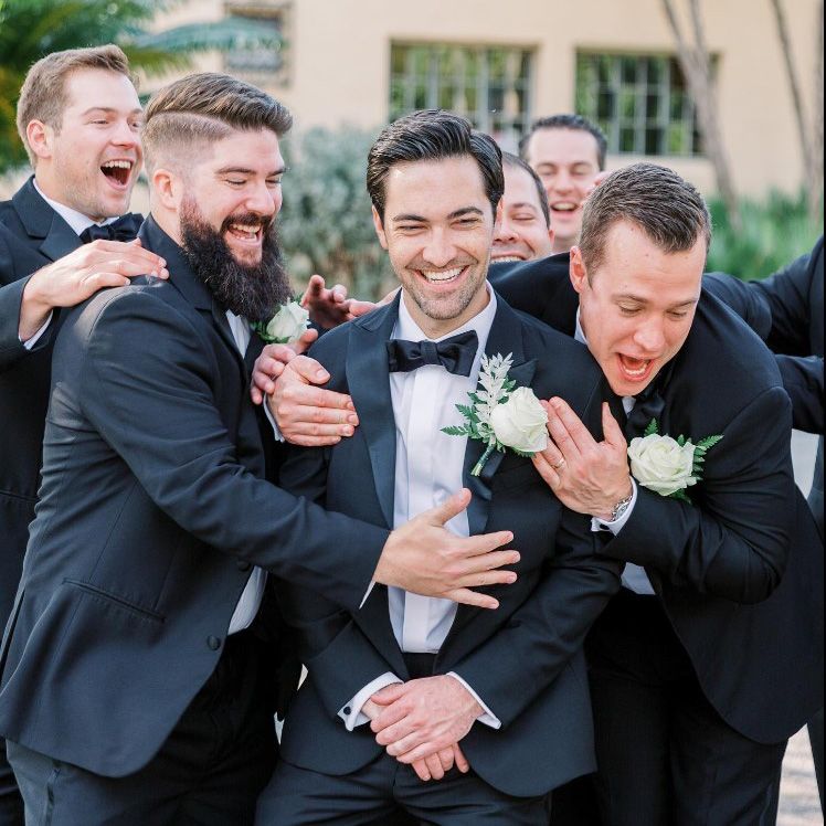 Image a groom with a white rose in his lapel laughing with 5 groomsmen surrounding him