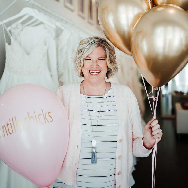 Woman laughing with blonde hair holding one pink balloon and a bunch of gold balloons in front a rack of white wedding gowns in a bucket with a lot of flowers in shades of pink, white and peach with a skyline view in the background