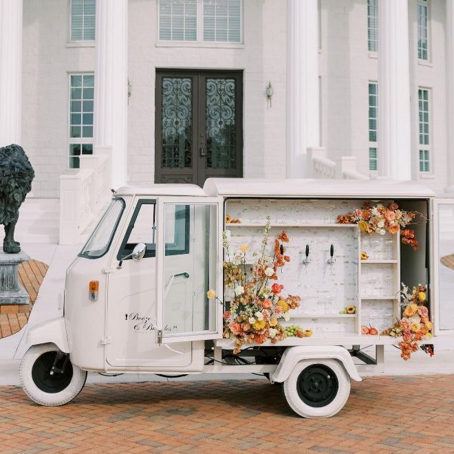 image of an old fashioned white truck with white wheels with taps on the side to service alcohol surrounded by splays of orange, yellow and white flowers with a white building with pillars and an ornate metal door in the background