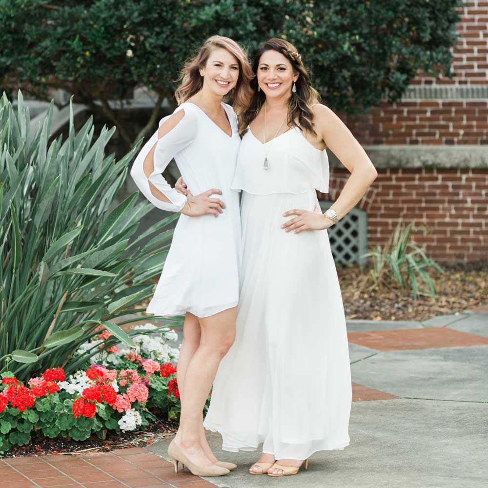 Photo of Confetti Events owners Krista and Kaylea wearing white and standing in a courtyard with red and white flowers