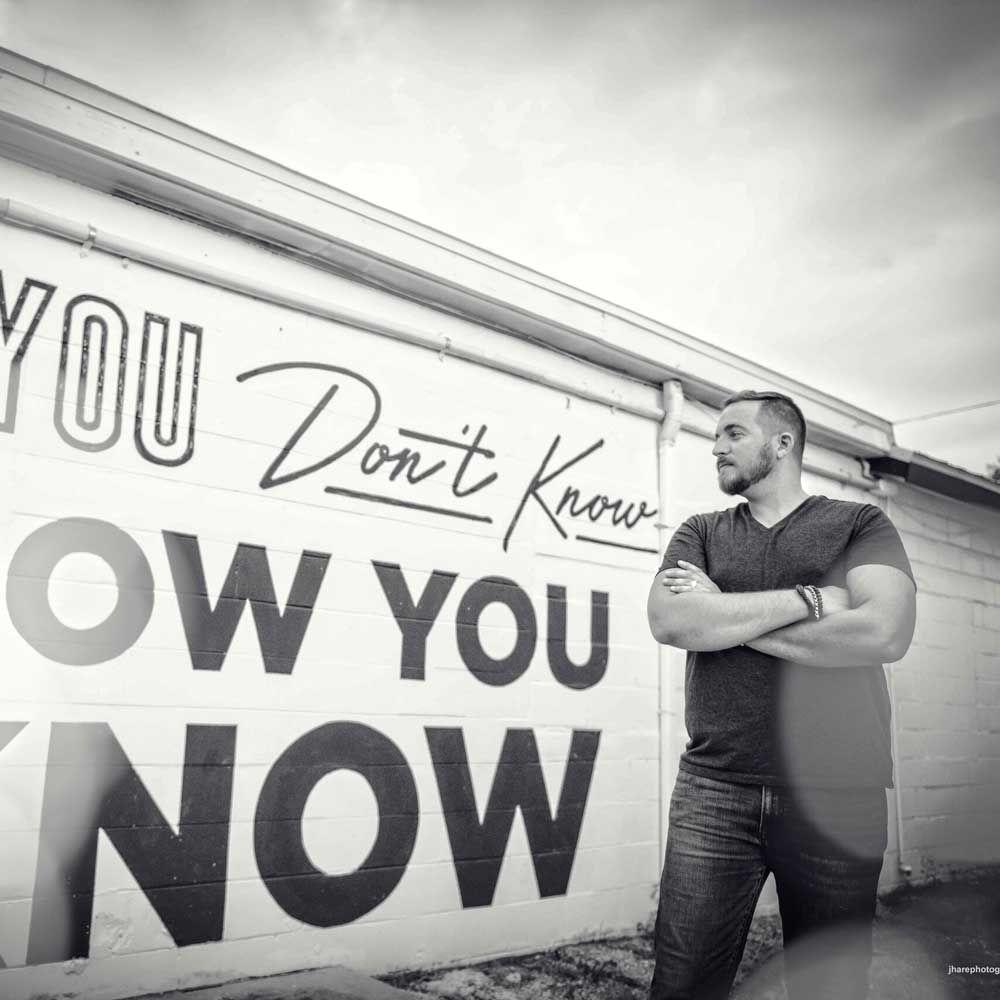 Image of Cory Barron standing next to graffiti that says "if you don't know, now you know."