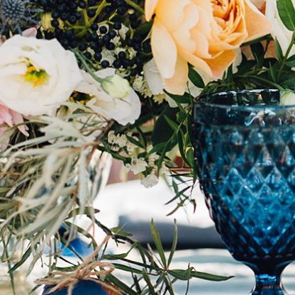 table setting with a dark blue glass goblet on a white tablecloth next to white and peach colored flowers