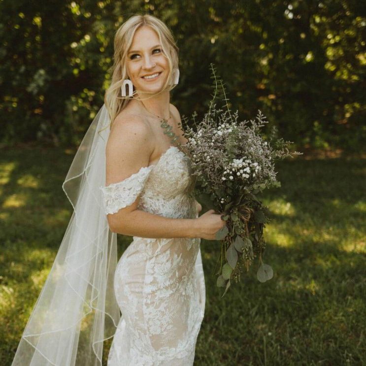 image of a smiling bride in a lace sleeveless dress holding a large bouquet with greenery and babies breath and a long train